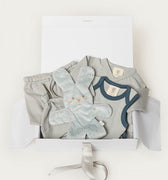 A baby gift set in a white box, featuring a light blue short-sleeve bodysuit with navy trim, a matching pair of light blue pants, a plush light blue bunny toy, and a white box with a gray ribbon.