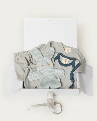 A baby gift set in a white box, featuring a light blue short-sleeve bodysuit with navy trim, a matching pair of light blue pants, a plush light blue bunny toy, and a white box with a gray ribbon.